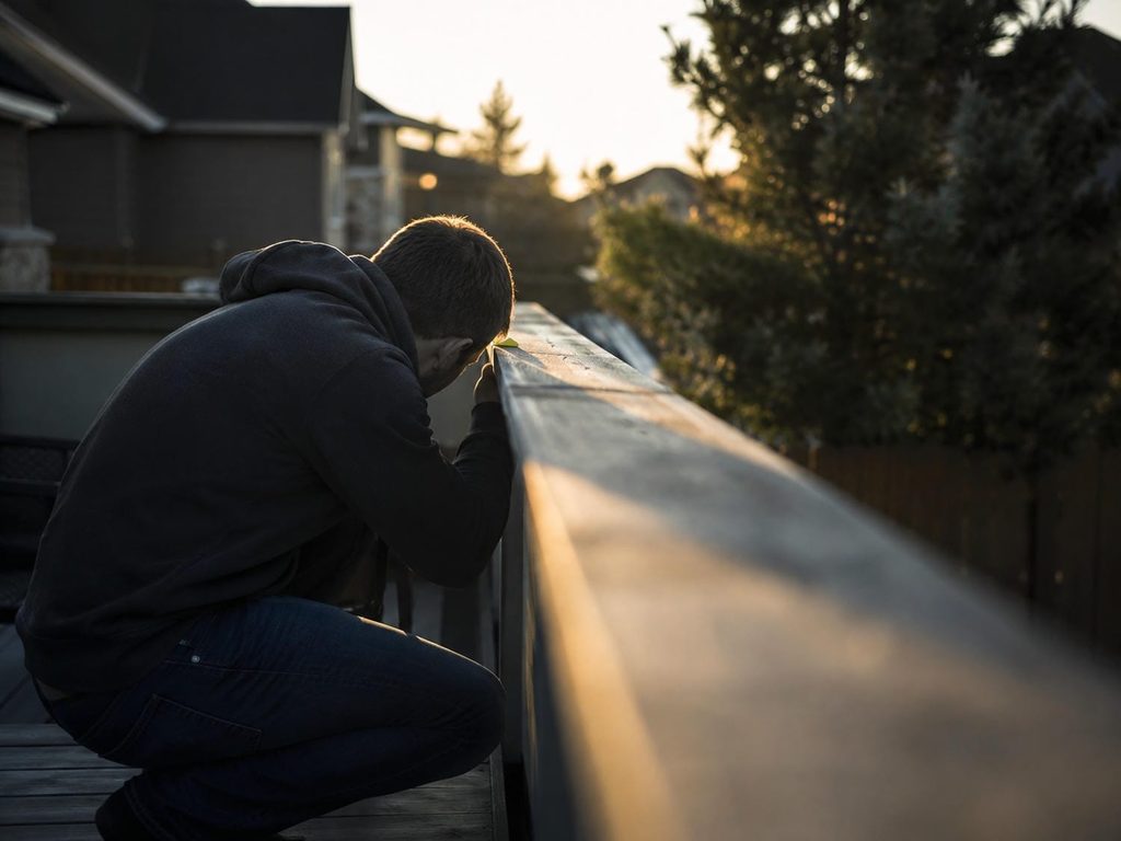Person crouched at deck level sighting along an aluminum railing in low sun to pick out fine lines under the coating