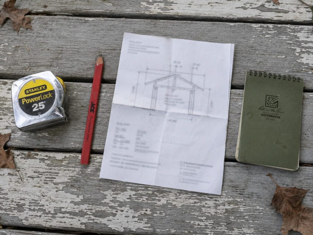 Tape measure, carpenter pencil and a folded drawing laid out on weathered porch floorboards